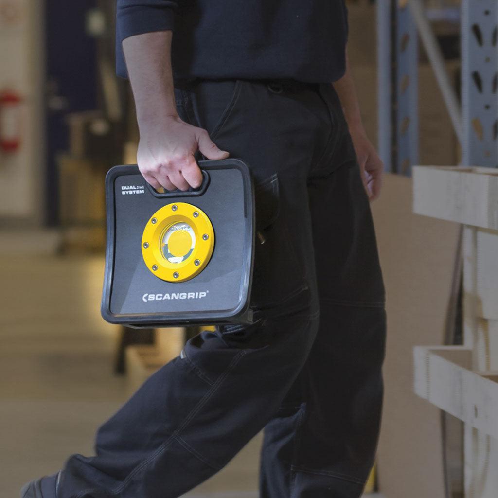 Persona sosteniendo foco LED portátil Scangrip negro y amarillo en ambiente industrial