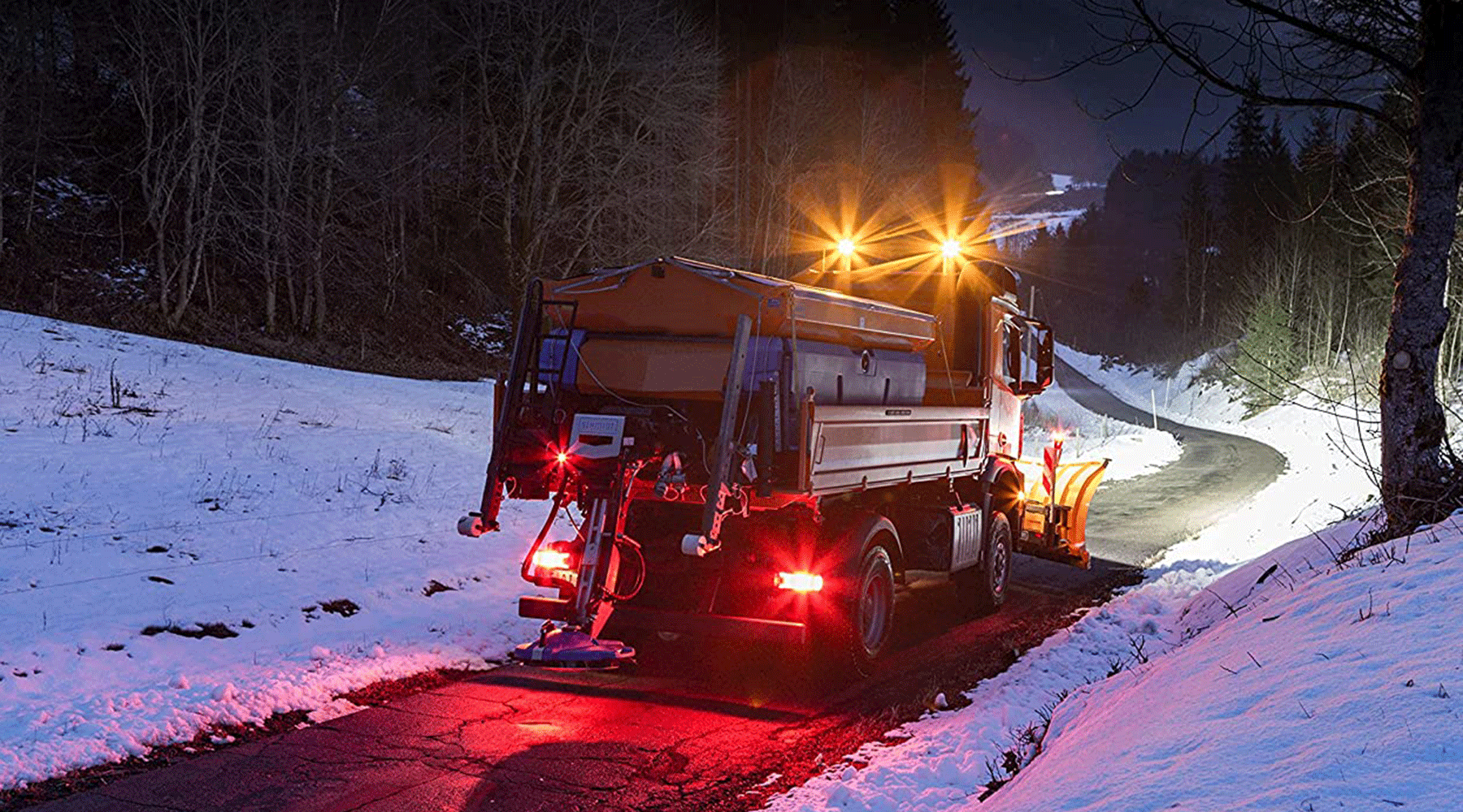 Camión quitanieves con luces LED encendidas limpiando carretera nevada de noche
